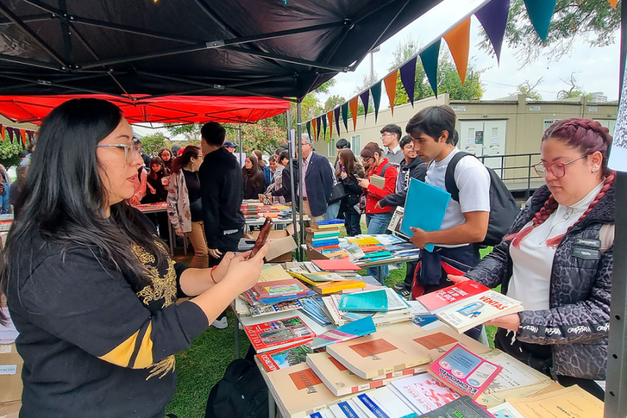 Funcionaria de la biblioteca de la Facultad de Humanidades entrega libros a la comunidad universitaria. En la fotografía se divisa un toldo y unas banderas triangulares de colores que cruzan el toldo.