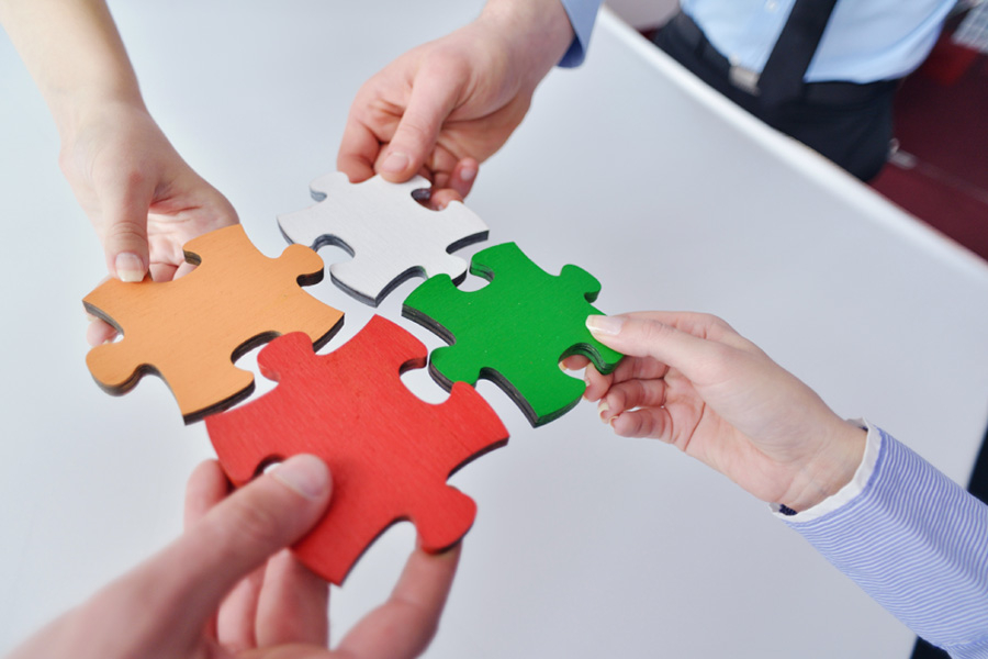 En la imagen aparecen 4 manos por separadas que intentan hacer calzar unas piezas de un rompecabezas. Las piezas son de colo amarillo, rojo, verde y blanco.