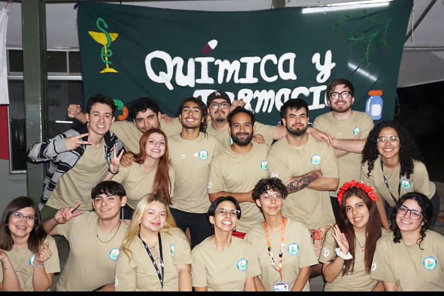 Estudiantes de la carrera de Química y Farmacia posan alegres a la fotografía. Levan una vestimenta café claro y en el fondo una bandera negra con letras blancas que enuncian el nombre de la carrera