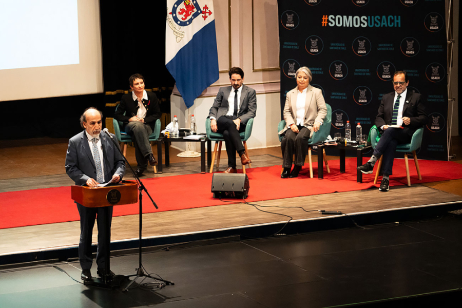 Candidatos y candidtas presidenciales del oficialismo sentados sobre unas sillas en el escenario del Teatro Aula Magna. Delante de ellos, el Rector Vidal da el inicio del Debate Presidencial.