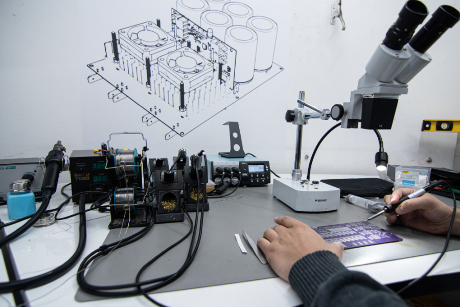 En la fotografía aparecen unas manos manipulando equipos del laboroatorio científico.  En el fondo, se observa un una muralla blanca y sobre el un dibujo 3D de un prototipo.