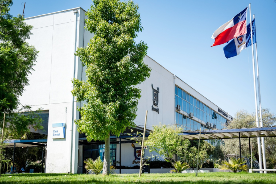 Frontis Casa Central Usach. En la fotografía aparecen las banderas de Chile y la universidad que flamean en dirección izquierda. También aparecen dos árboles que acompañan la fachada. 