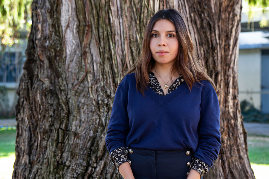 En la fotografía aparece la investigadora Valeria Olivares. Viste una chaleco azul oscuro con escote en V, camisa negra con vivos blancos y pantalones negros.Detrás suyo el tronco de un árbol. 