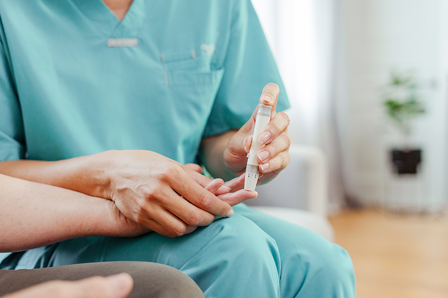 Imagem desfocada de um profissional de saúde com bata turquesa segurando um dedo e um dispositivo médico, possivelmente uma lanceta, para medir o nível de açúcar no sangue.