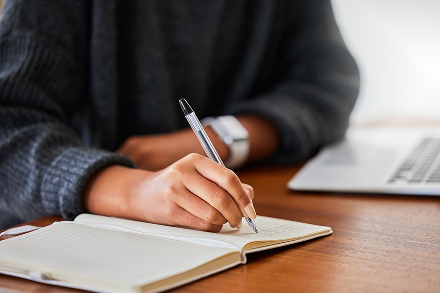 Se muestran las manos de una persona escribiendo en un cuaderno sobre un escritorio de madera. Junto al cuaderno hay un computador.