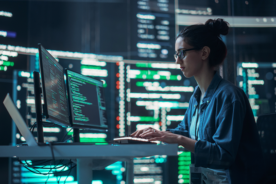 A focused woman wearing glasses is working on a computer at a desk in a dark room. The screens behind her display many lines of code and data.