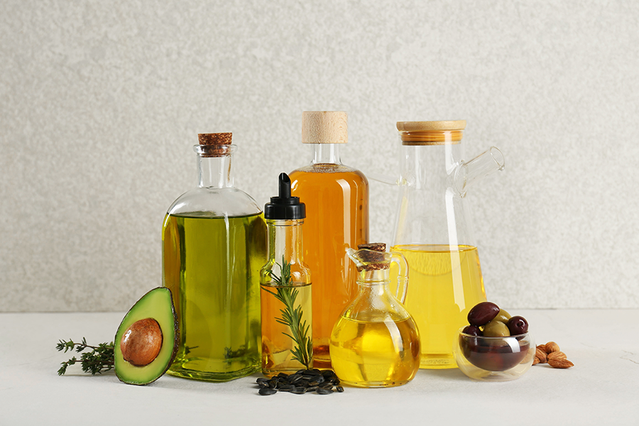 A variety of oils are displayed in different glass bottles, along with a halved avocado, olives, and almonds, on a light-colored surface.