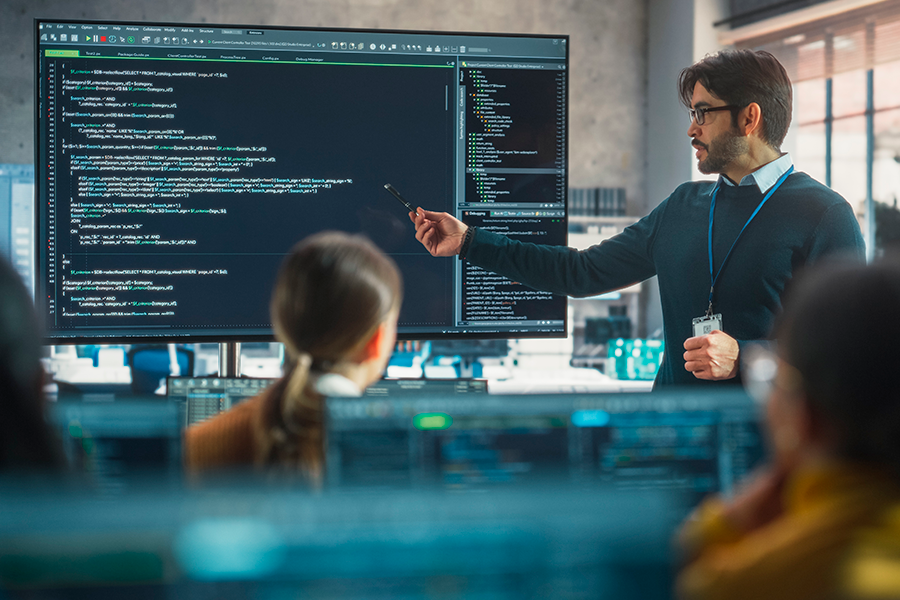 Un profesor señala una gran pantalla en la que se muestran líneas de código informático durante una clase en un entorno educativo tecnológico.