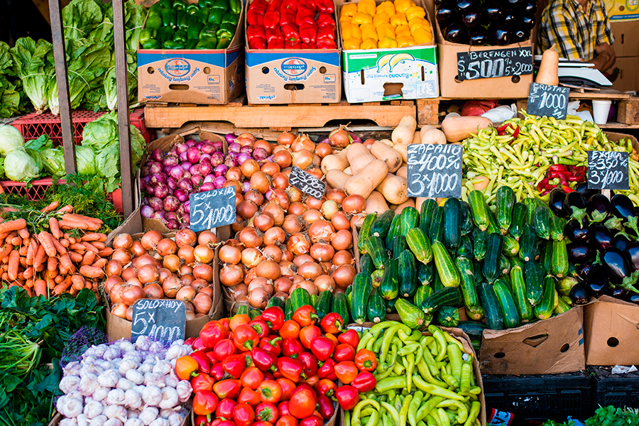 Esta fotografía captura una vibrante y abundante muestra de productos frescos en un mercado callejero, con montones de pimientos, cebollas, zanahorias, calabazas, ajos y pepinos de colores, muchos de ellos con etiquetas de precios escritas a mano.