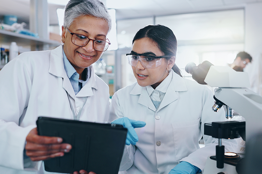Two scientists, one older with glasses and the other younger with safety glasses, collaborate and look at a tablet together in a laboratory.