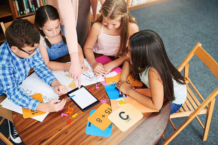 Un docente señala un tablet mientras cuatro estudiantes trabajan en actividades matemáticas y de aprendizaje alrededor de una mesa en un aula.