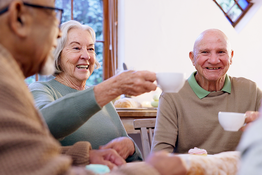 Três idosos sorridentes apreciam uma xícara de chá ao redor de uma mesa, sob uma luz acolhedora, e um deles ri enquanto segura sua xícara.