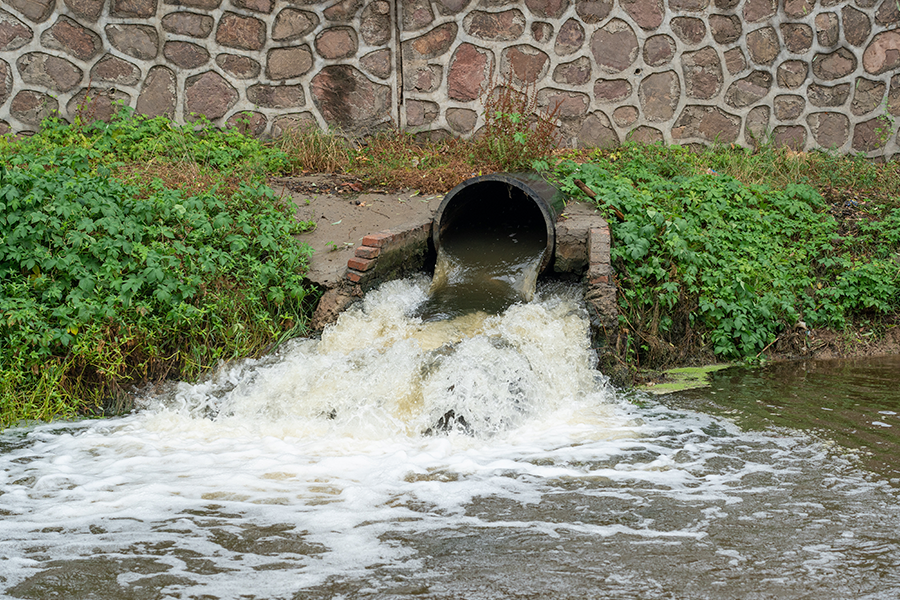 Um tubo preto emerge de uma parede de pedra, liberando um jato potente de água espumosa e lamacenta em um rio turvo, rodeado por algas verdes.
