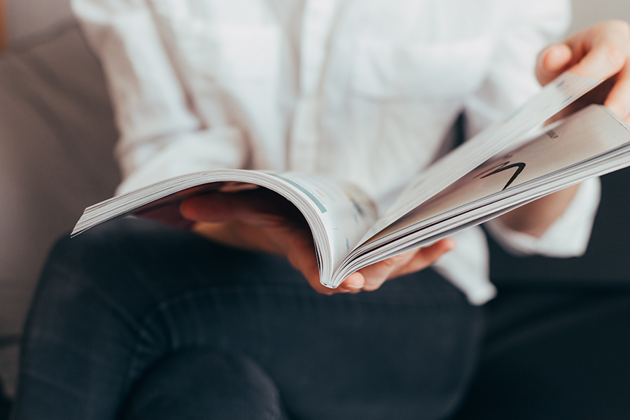 Una persona con camisa blanca y pantalones oscuros, sentada, hojea las páginas de una revista, con las manos sujetando el lomo y pasando las páginas.