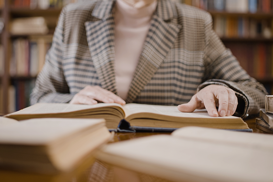 A person in a plaid blazer sits at a desk in a library, their hands resting on the pages of a large, open hardcover book.