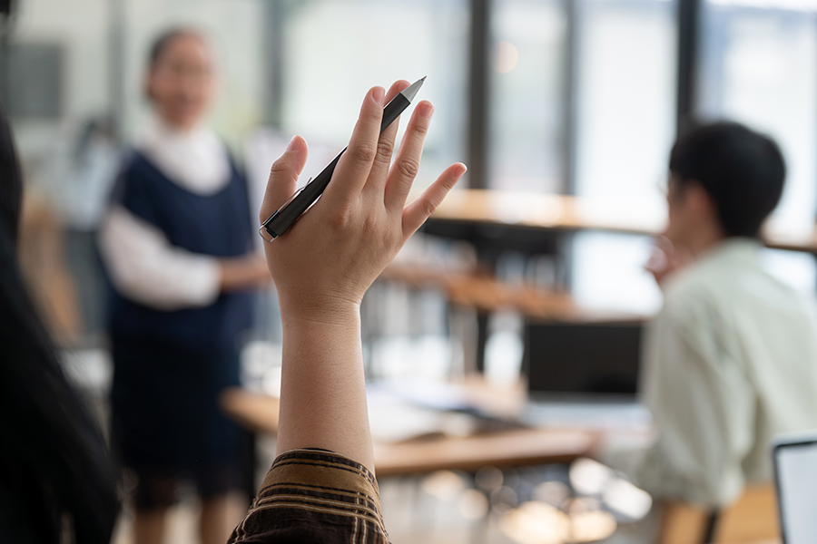 A person's hand holds a black pen high in the air to ask a question during a classroom presentation or lecture.