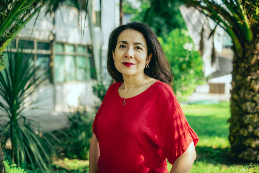 En la fotografía aparece la Dra. Claudia Vargas con un vestido rojo. De fondo, los patios de la Usach