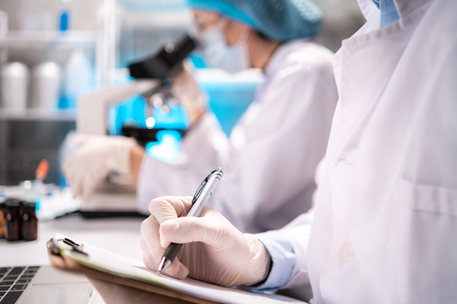 Scientists in white coats and blue caps work in a lab, focusing on microscopes and taking notes.