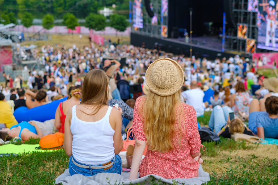 Back view of two women sitting on grass at an outdoor festival with a large stage in the distance.
