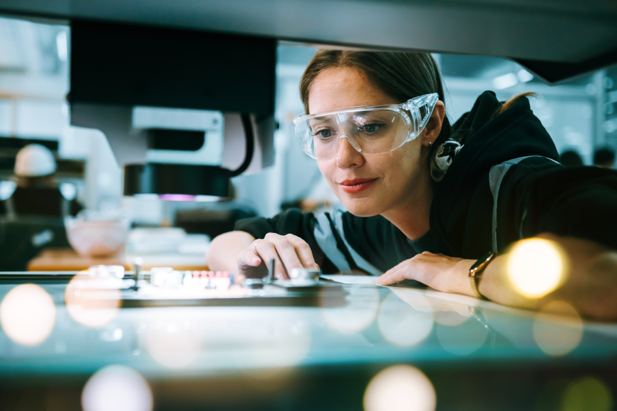 Mujer con gafas de seguridad mirando de cerca un espacio de trabajo de cristal iluminado y resplandeciente.