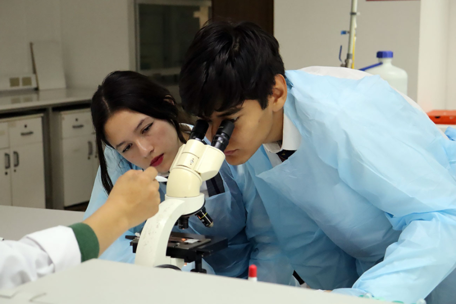 La imagen muestra a dos estudiantes en un laboratorio, un hombre y una mujer. El hombre está mirando por el microscopio. Ambos usan un delantal celeste