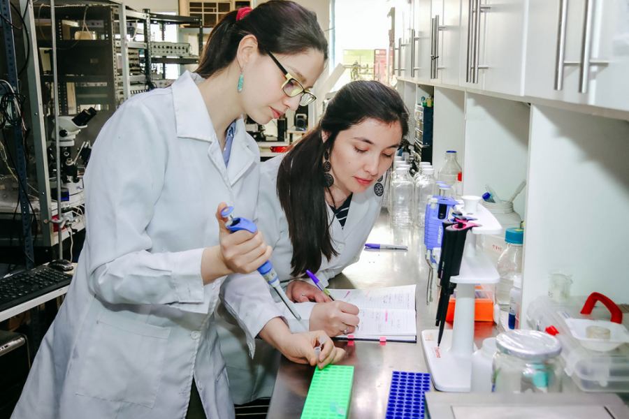 La imagen muestra a dos mujeres científicas con delantal blanco en un laboratorio: una manipulando instrumentos y la otra registrando lo observado