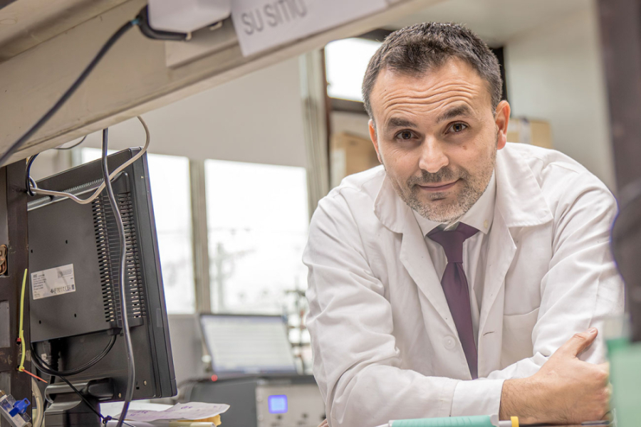 La foto muestra al Dr. Jaime Pizarro apoyado sobre una mesa, con cotona blanca y corbata morada, en su laboratorio
