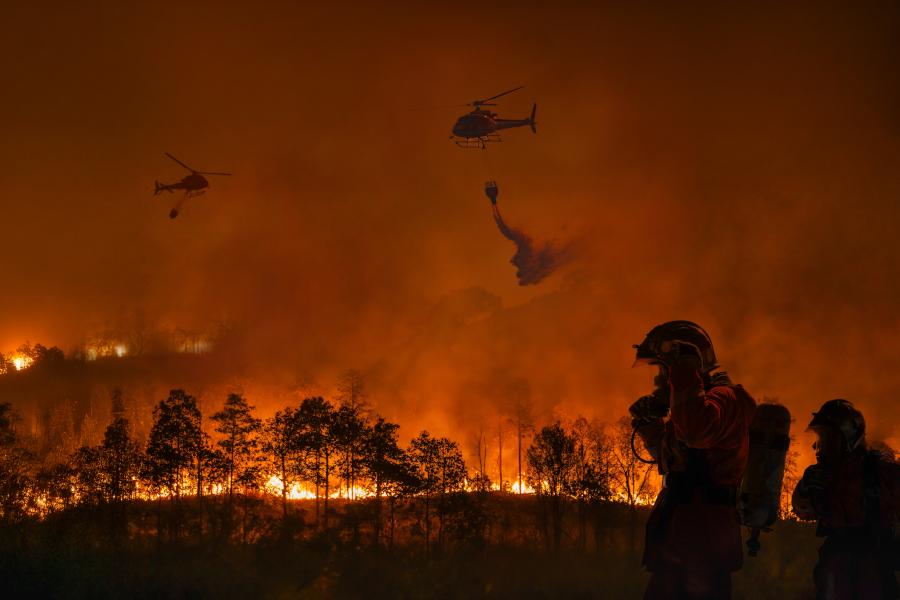 Firefighters battle a massive forest fire as helicopters drop water.