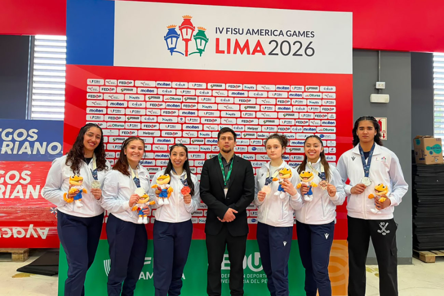 La imagen muestra a las estudiantes posando con sus medallas y un peluche de mascota de los juegos. Al medio su entrenador y de fondo el pendón del evento