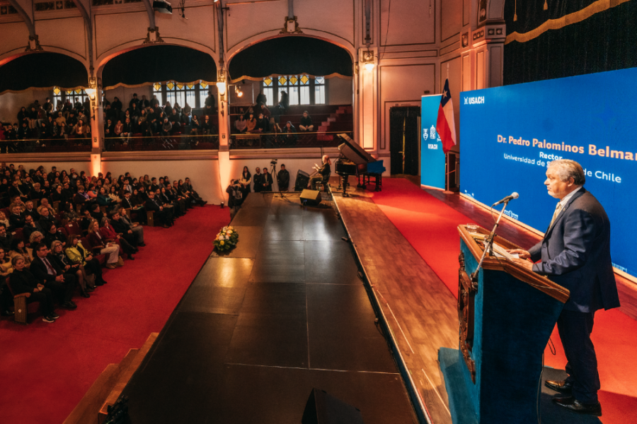 La imagen muestra la ceremonia de aniversario en el Aula Magna. En la esquina inferior derecha aparece el rector en el podio dando un discurso y al lado izquierdo de la imagen, el público