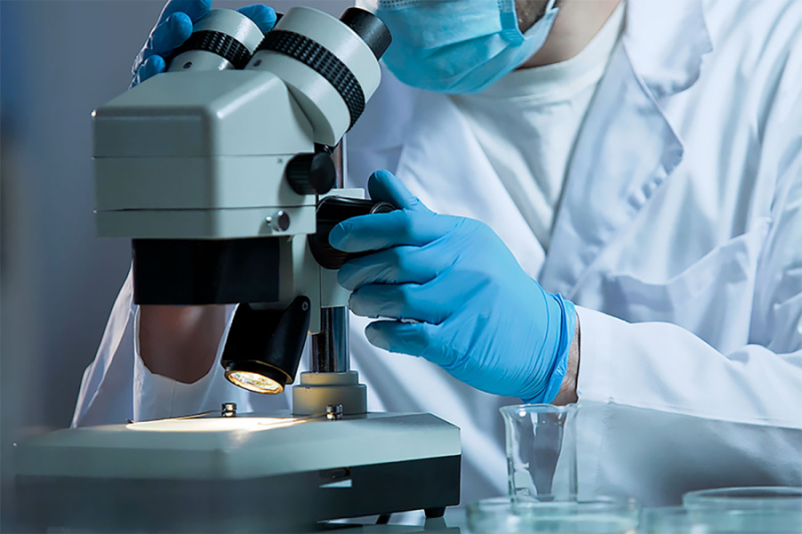 En la imagen aparece un científico trabajando sobre un microscopio. Aparece con delantal blanco y guantes celestes.
